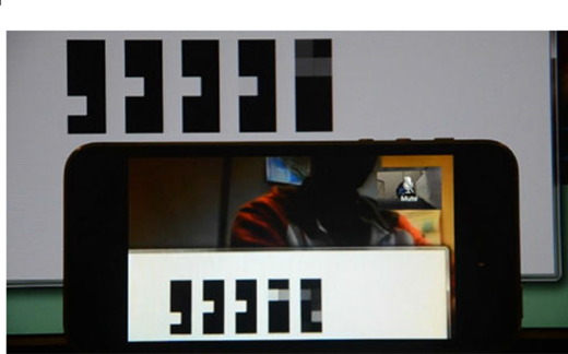 A photograph showing an automated latency measurement setup, where a smartphone displays a real-time video of a PC monitor, both showing synchronized binary-based visual markers to calculate end-to-end delay.