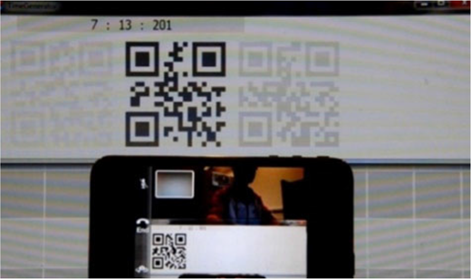 A photograph showing a technical setup to measure video latency, where a smartphone captures a PC monitor’s screen displaying a digital timestamp 7 : 13 : 201 and a large QR code.