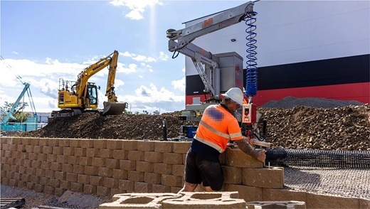 A construction worker and robotic equipment operate on a brick wall at an active building site.