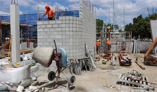 Several construction workers build a concrete block wall on a large construction site using scaffolding and masonry tools.