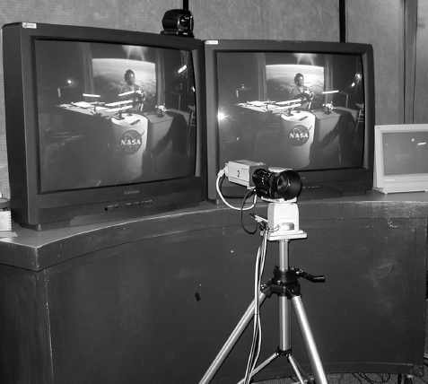 A photograph showing two television monitors displaying the same video feed of an astronaut in space (or a simulation thereof) sitting at a desk with a N A S A logo.