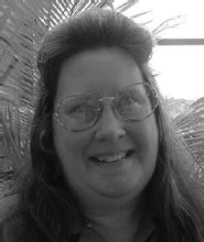 A closeup photograph of a woman with glasses and long hair, smiling at the viewer with foliage in the background.