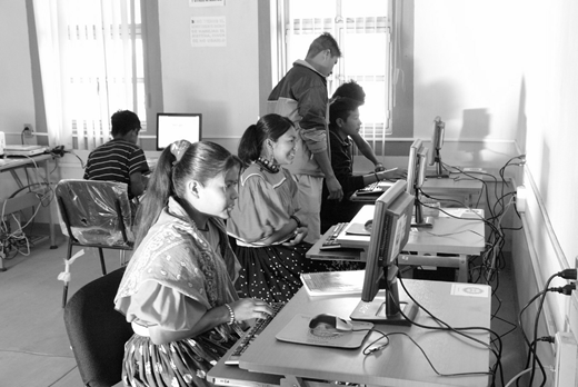 A photograph shows four students, two females in the foreground and two males in the background, seated at computers in a classroom, with one male student standing between the seated students.