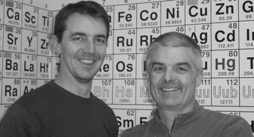 A photograph shows two males smiling at the camera in front of a wall covered with a Periodic Table of Elements.