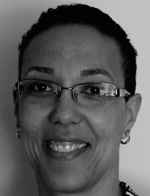Black and white portrait of a person with short hair and glasses, smiling warmly. They wear hoop earrings, conveying a friendly and approachable tone.