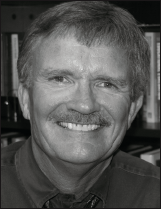 A smiling man with glasses and graying hair, wearing a striped shirt, is pictured in this black and white portrait. The tone is warm and approachable.