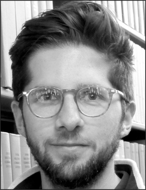 A librarian with short hair sits in front of bookshelf containing multiple books and documents.
