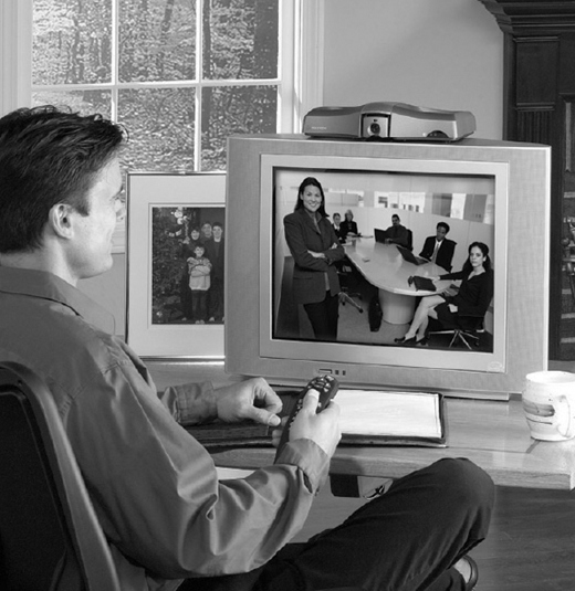 A man in a room, watching a video conference on a television set, holding a remote control.