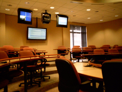 A photo shows a modern lecture hall with tiered curved desks, many swivel chairs, wallmounted windows, and four overhead screens including monitors and a projector.