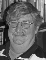 Black and white portrait of a smiling man with glasses and a mustache, set against a backdrop of bookshelves. The tone is warm and approachable.