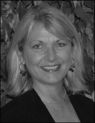 Black-and-white portrait of a smiling middle-aged woman with shoulder-length light-colored hair parted to the side. She is facing the camera with a warm, friendly expression, wearing subtle earrings, a delicate necklace, and a dark top or blazer. The background features softly focused foliage or textured leaves, adding depth and a natural feel to the image. The lighting highlights her facial features and smile, giving the photo a professional yet approachable look.