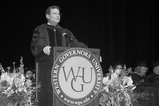 A grayscale photo of a speaker in academic regalia standing at a podium with the Western Governors University seal, surrounded by floral arrangements and seated individuals in the background.