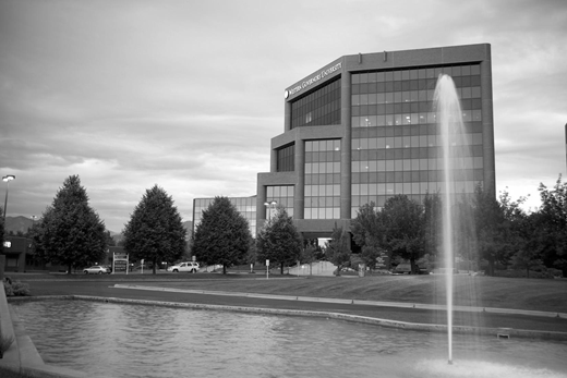 A grayscale photo of a mid-rise office building with large windows, a row of trees in front, and a tall fountain spraying water in the foreground near a pond.