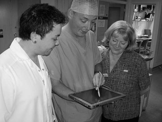 A grayscale photo of three medical staff standing together while one uses a stylus on a tablet device as the others watch.