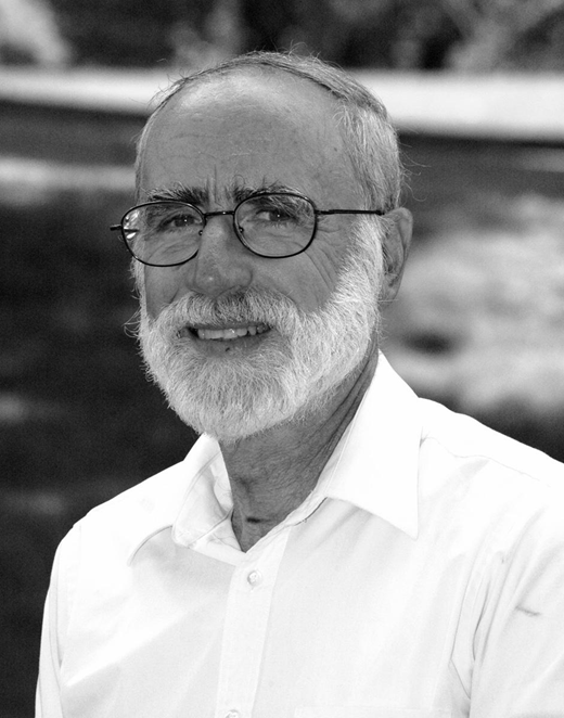 Portrait of an older man with glasses and a beard, wearing a light shirt and standing outdoors.