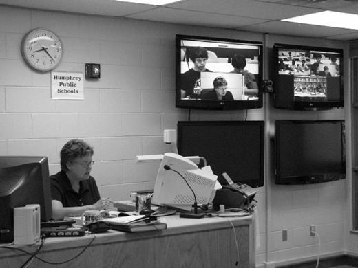 Person at desk in school control room monitors classroom video feeds on wall screens labeled Humphrey Public Schools; two screens show live footage, others are off; desk holds office equipment.