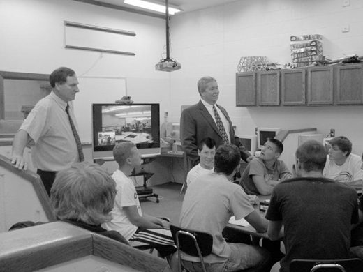 Students sit around a table in a tech-equipped classroom as two men lead a discussion; a monitor shows a mirrored room view, with computers, projector, and supplies visible in the background.