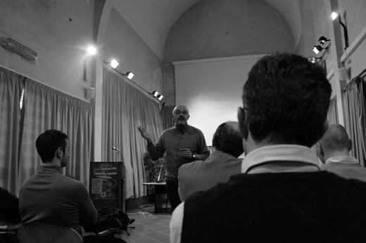 Black and white photo of a man presenting in a room with arched ceilings, projection screen, and seated audience. A podium sign reads Associazione Culturale L'Orto degli Ulivi.