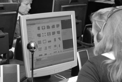 Black and white photo of a person wearing headphones at a computer with webcam and editing software; others work at nearby stations in a tech-focused lab or office setting.