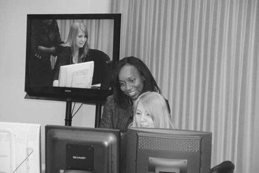 Black and white photo of two people at computer monitors, with a TV screen behind showing their live video feed in a lab or office setting.