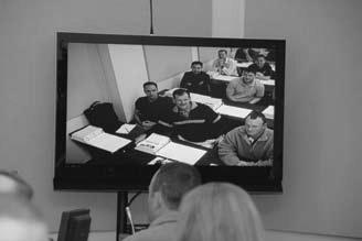 Black and white photo of a TV screen showing students seated at desks in a classroom, viewed by people in the foreground, suggesting a remote learning or video-based educational setup.