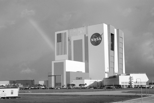 A black and white photo of the NASA Vehicle Assembly Building at Kennedy Space Center, with a large US flag and NASA logo.