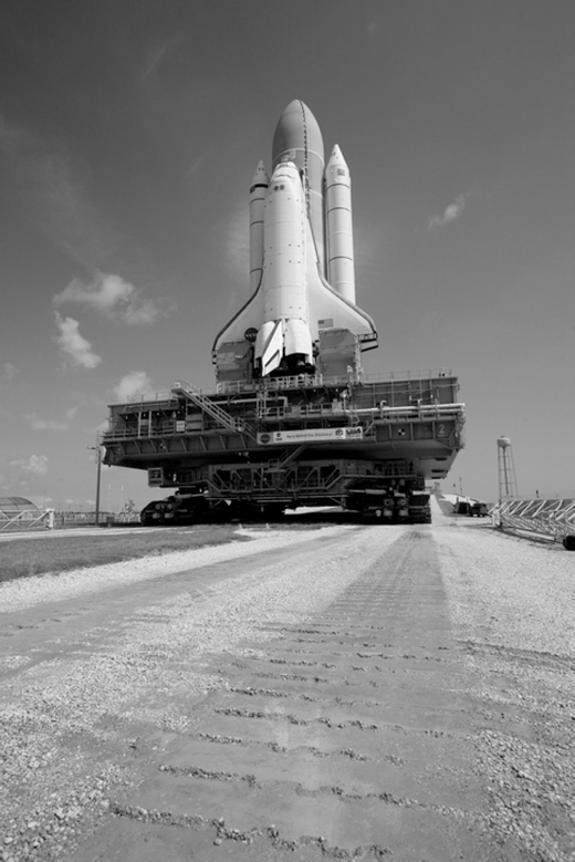 A black and white photo of a Space Shuttle stack on the Crawler-Transporter, moving along the gravel path.