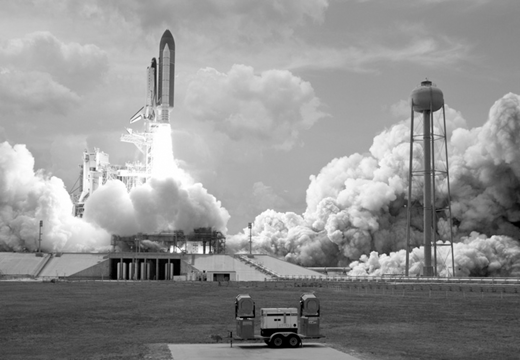 A black and white photo of the Space Shuttle STS-125 launching from the pad, surrounded by fire and smoke.