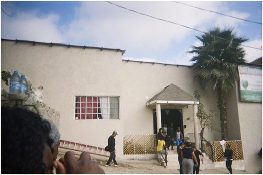 A photograph of a building with people near the entrance, a palm tree on the right, and water bottles on a rock wall.