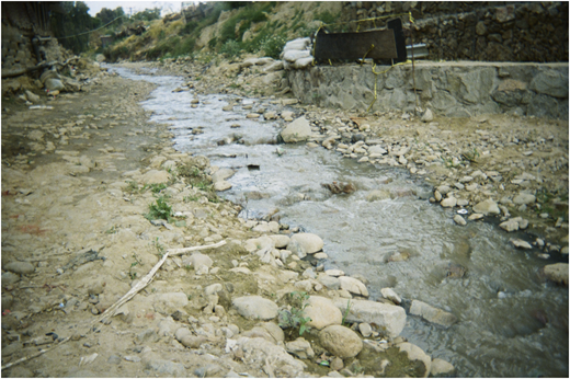 A photograph of a shallow rocky stream flowing between dirt banks with scattered stones and a stone wall.