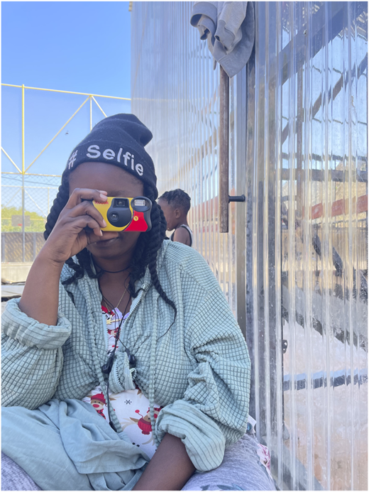 A photograph of a person holding a small camera while seated against a clear paneled wall.