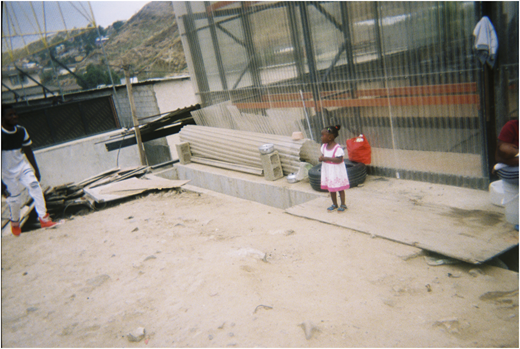 A photograph of a child in a pink dress standing near a wall with stacked materials, with people and debris around.