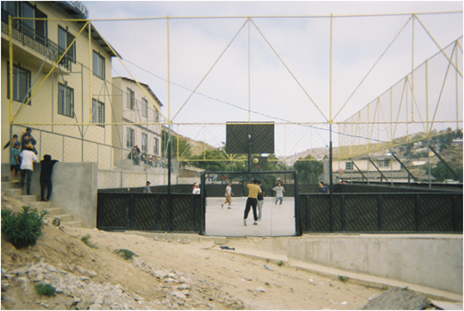 A photograph of people on an outdoor court with a tall fence and children on nearby steps.
