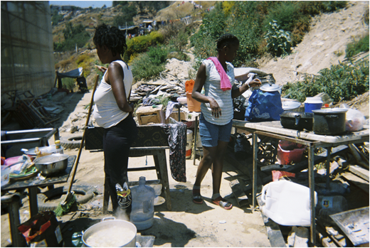 A photograph of two people working at outdoor tables covered with cooking pots and supplies on a dirt slope.