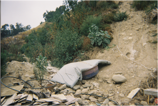 A photograph of a dirt hillside with bushes, rocks, and a covered round object near loose boards and cables.