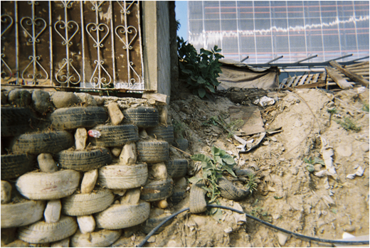 A photograph of a wall made of stacked tires beside a metal gate, with dirt, plants, and scattered boards on a slope.