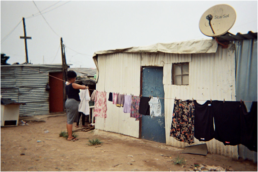 A photograph of a woman hanging clothes on a line beside corrugated metal structures with a door, window, and satellite dish.