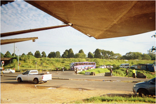 A roadside scene with vehicles, pedestrians, traffic lights, and a bus beside a grassy area under a large canopy.