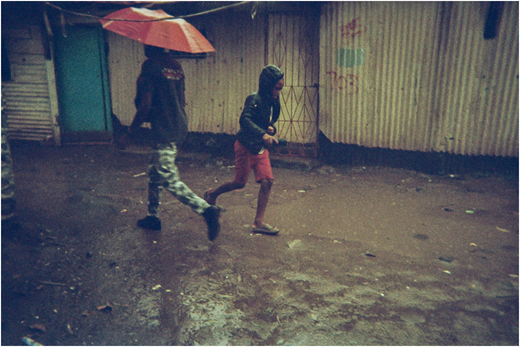 A photograph of two people moving through a muddy open area beside corrugated metal walls, one holding an umbrella.
