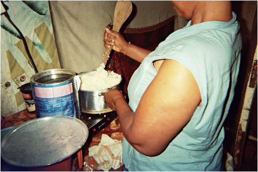 A photograph of a person stirring food in a pot on a small stove beside cans, a lid, and fabric-covered surfaces.