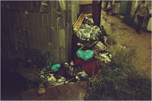 A photograph of a pile of discarded bottles and bags beside metal walls, with grass and a narrow muddy walkway nearby.
