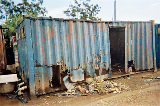 A photograph of a rusted blue metal container with holes, an open doorway, scattered debris, and mud on the ground.