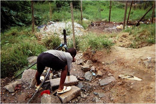 A photograph of a person bending near a water outlet among rocks and debris, with grass and a wire fence in the background.