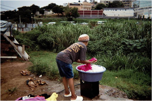 A photograph of a person washing clothes outdoors beside a large basin, with grass, debris, and buildings in the background.