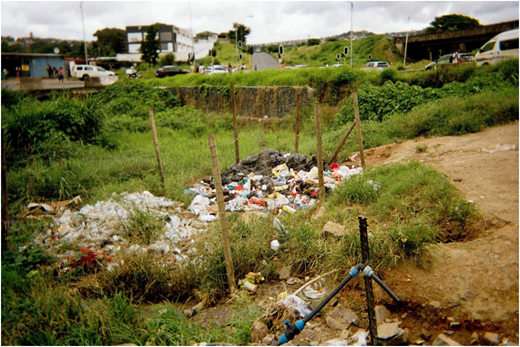 A photograph of a grassy area with piles of trash behind a wire fence, with roads, vehicles, and buildings in the background.
