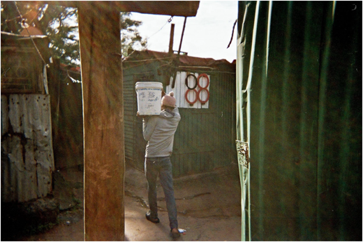 A photograph of a person carrying a bucket through an outdoor passage between metal structures.