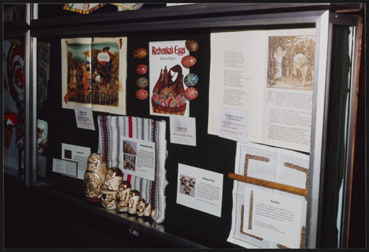 A display case presents illustrated book pages, decorated eggs, nesting dolls, a woven textile, a wooden flute, and printed information cards arranged together.