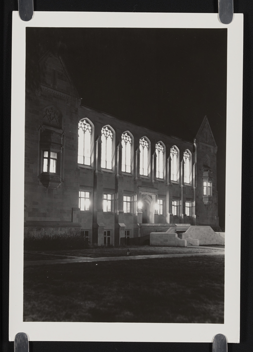 A large stone building with tall arched windows and a central entrance is photographed at night, with illuminated windows and steps leading to the doorway.