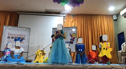 A stage scene showing children in themed costumes, with one child speaking at a microphone and a banner reading Bienvenidos al 3 Festival De Fantasía in the background.