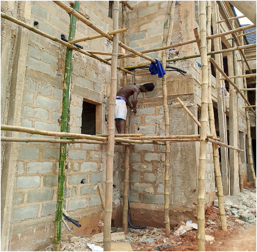 A photograph of a carpenter standing on bamboo scaffolding beside a brick building wall, working without protective gear.
